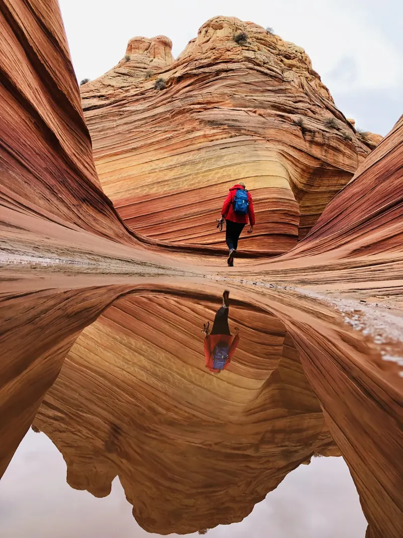 A hiker in a red jacket walking through The Wave, reflected in a rain pool